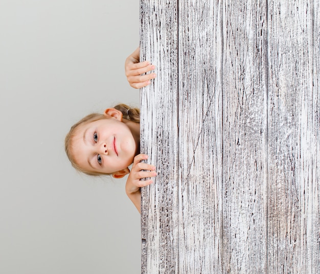 Little girl hiding behind wooden plank and looking confident .