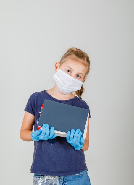 Little girl holding books and notebooks in t-shirt, gloves and medical mask front view.