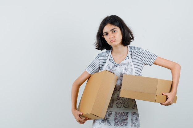 Little girl holding cardboard boxes in t-shirt, apron and looking doubtful.