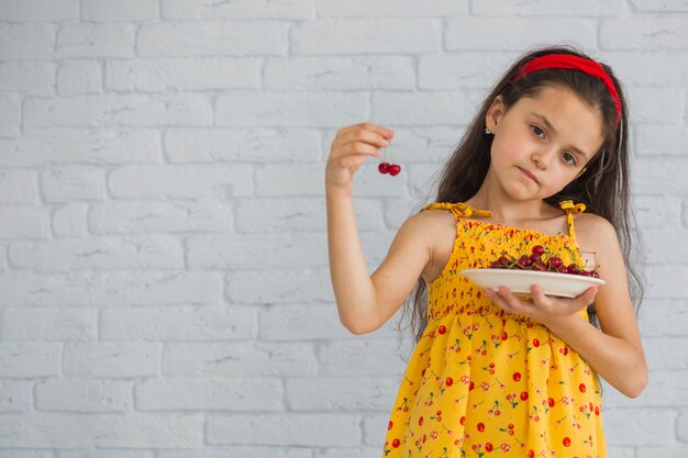Little girl holding cherry and plate in front of white brick wall