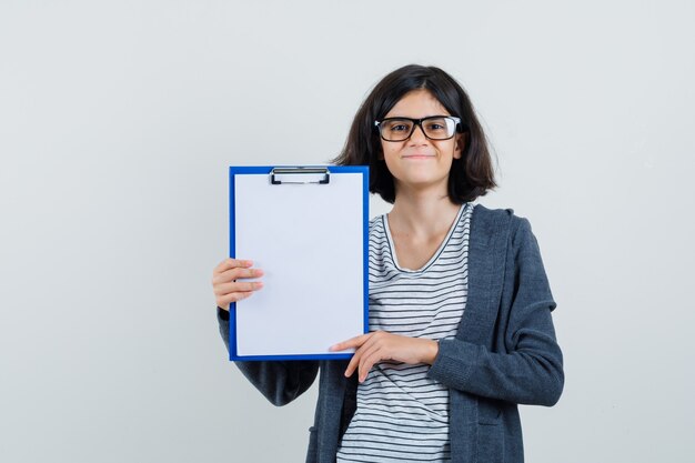 Little girl holding clipboard in t-shirt, jacket and looking cheery.