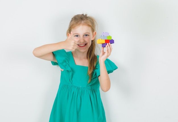 Маленькая девочка держит красочные блоки конструктора с большим пальцем руки вверх в зеленом платье и выглядит веселой. передний план.