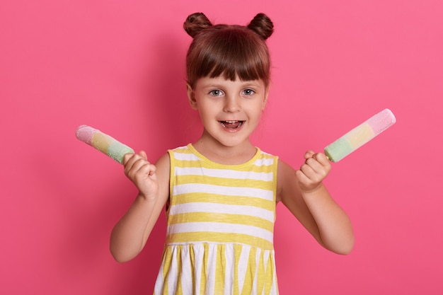 Little girl holding colorful ice lollies, having excited facial expression, posing with happy look, wearing white and yellow stripped summer dress against pink wall.