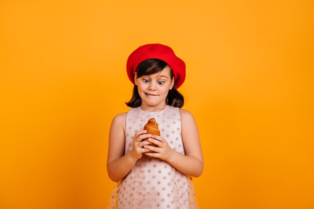 little girl holding croissant. Brunette kid in french beret expressing amazement on yellow wall.