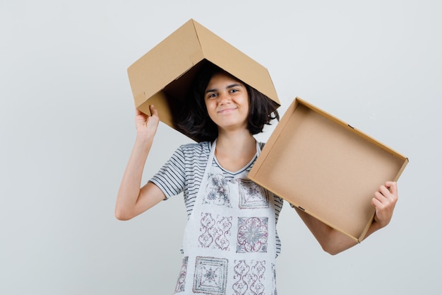 Little girl holding empty cardboard box in t-shirt, apron and looking cheery ,