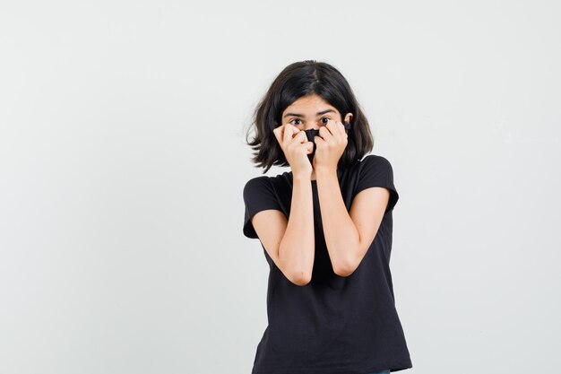 Little girl holding fists on face in black t-shirt, mask and looking scared , front view.