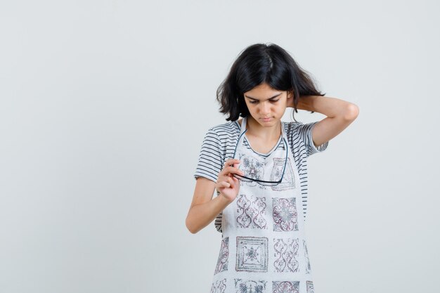 Little girl holding glasses, scratching head in t-shirt, apron and looking pensive ,