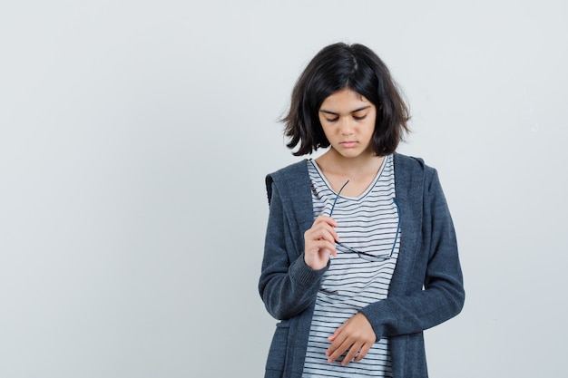 Little girl holding glasses in t-shirt, jacket and looking depressed.