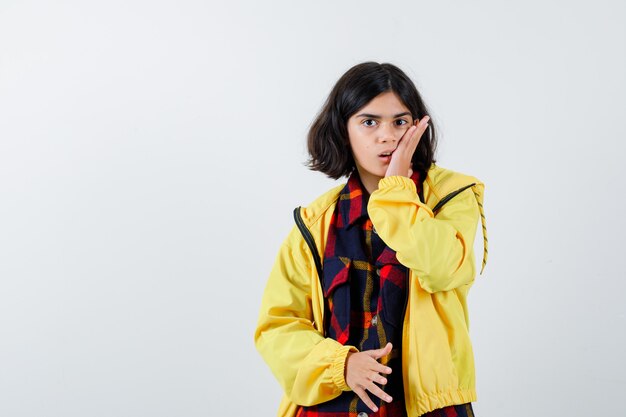 Little girl holding hand on cheek in checked shirt, jacket and looking amazed 