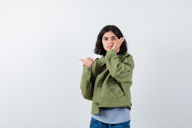 Little girl holding hand on cheek, pointing aside with thumb in coat, t-shirt, jeans and looking amazed , front view.