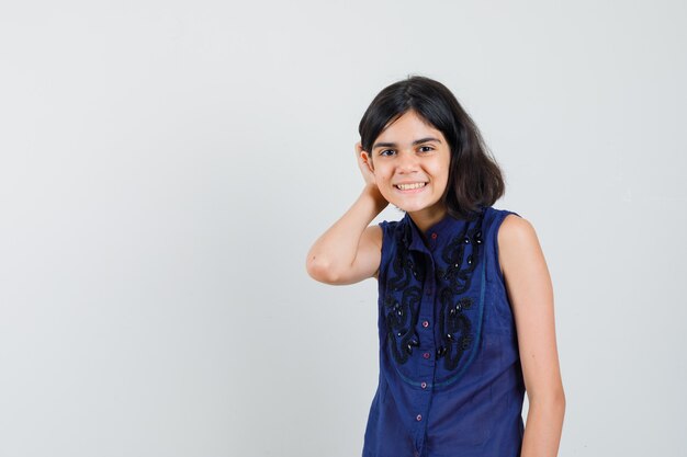 Little girl holding hand behind ear in blue blouse and looking glad. front view.