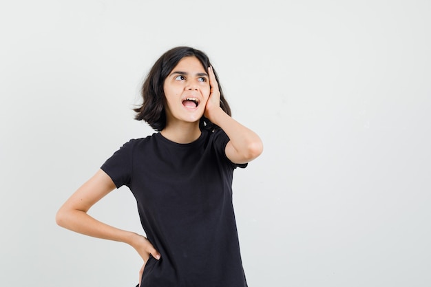 Little girl holding hand on face in black t-shirt and looking forgetful , front view.