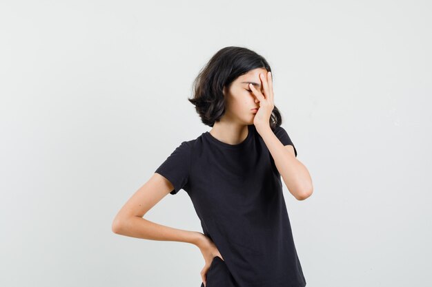 Little girl holding hand on face in black t-shirt and looking sleepy , front view.