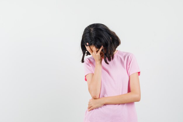 Маленькая девочка держит руку на лице в розовой футболке и смотрит задумчиво, вид спереди.