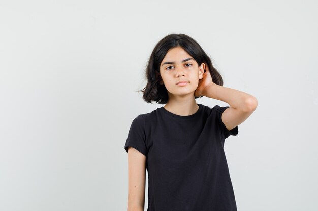Маленькая девочка держит руку в волосах в черной футболке и выглядит серьезным, вид спереди.