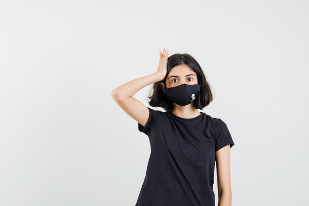 Little girl holding hand to head in black t-shirt, mask and looking wistful , front view.