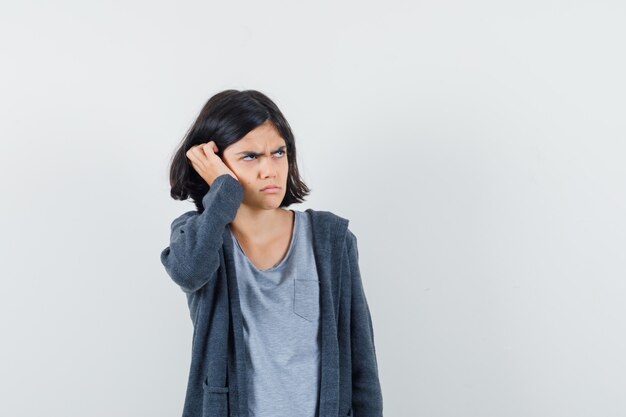 Little girl holding hand on head in t-shirt, jacket and looking upset