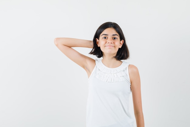 Little girl holding hand behind head in white blouse and looking merry. front view.