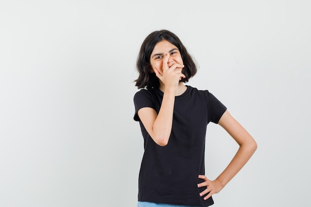 Little girl holding hand on mouth in black t-shirt and looking happy. front view.