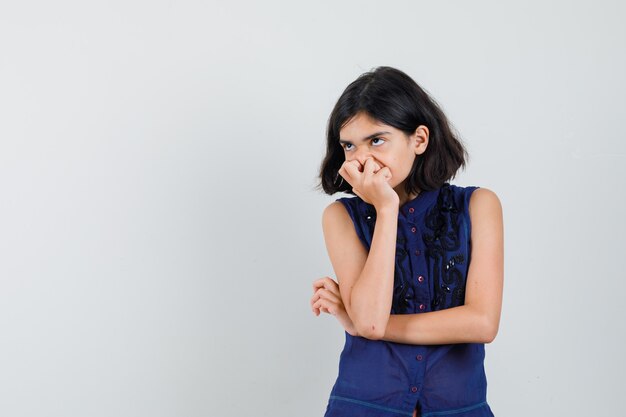 Little girl holding hand on mouth in blue blouse and looking pensive. front view.