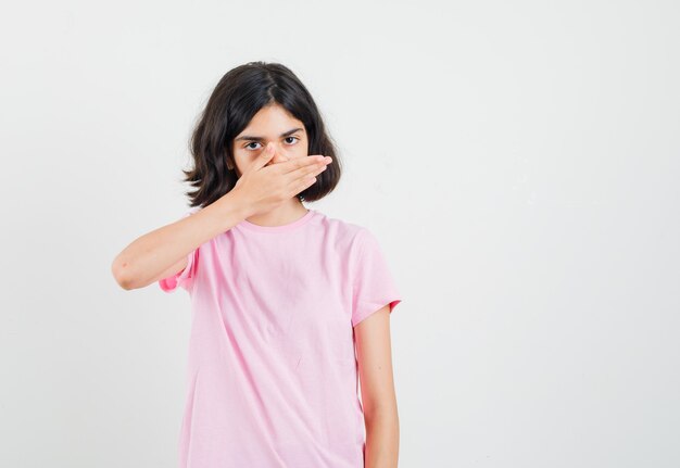 Little girl holding hand on mouth in pink t-shirt and looking scared. front view.