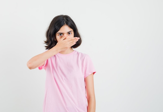 Маленькая девочка держит руку на рту в розовой футболке и выглядит испуганной. передний план.