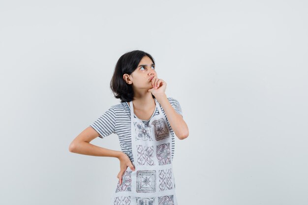 Little girl holding hand on mouth in t-shirt, apron and looking hesitant ,