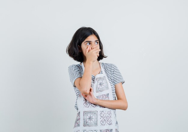 Little girl holding hand on mouth in t-shirt, apron and looking surprised.