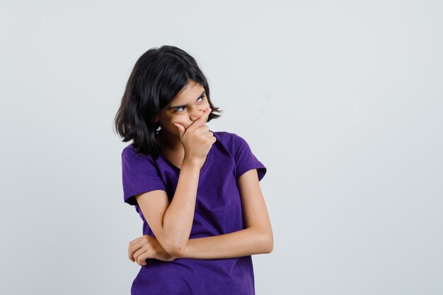 Little girl holding hand on mouth in t-shirt and looking pensive.