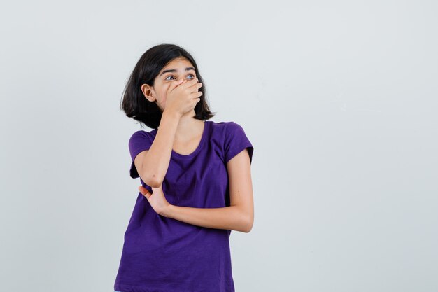 Little girl holding hand on mouth in t-shirt and looking surprised.