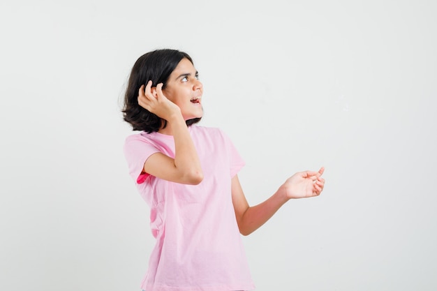 Little girl holding hand near ear in pink t-shirt and looking curious. front view.