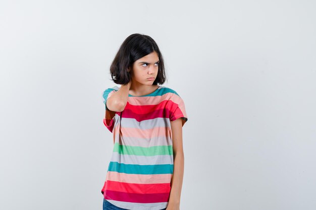 Little girl holding hand on neck, looking away in t-shirt and looking displeased , front view.