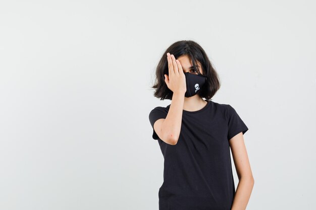 Little girl holding hand on one eye in black t-shirt, mask front view.