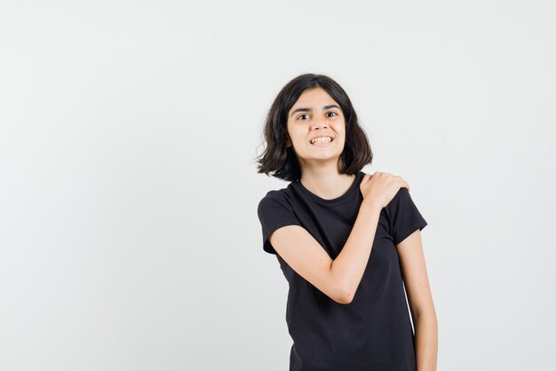 Little girl holding hand on shoulder in black t-shirt and looking glad , front view.