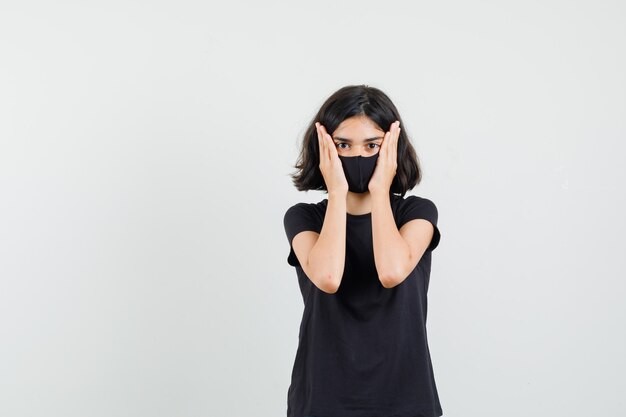 Little girl holding hands on cheeks in black t-shirt, mask front view.