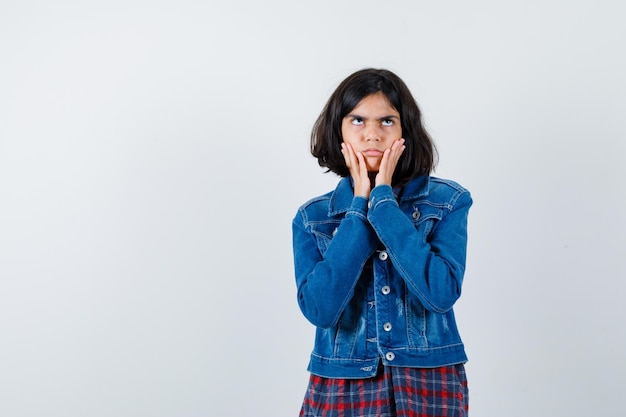 Little girl holding hands on cheeks in shirt, jacket and looking hesitant. front view.