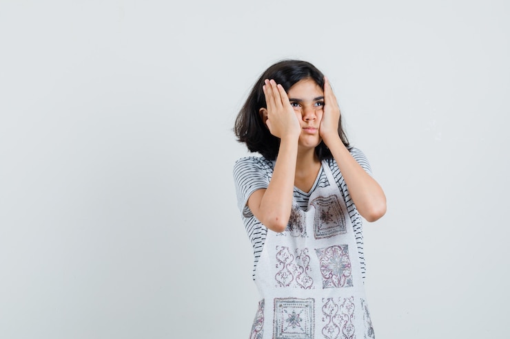 मुफ़्त फोटो little girl holding hands on cheeks in t-shirt, apron and looking wistful ,