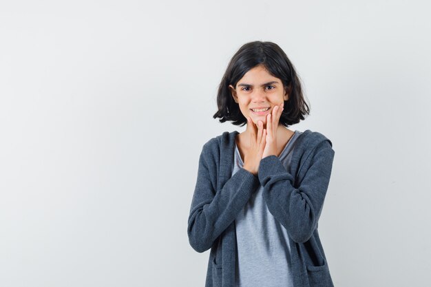 Little girl holding hands on chin in t-shirt, jacket and looking excited.