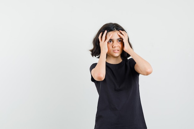 Little girl holding hands to face in black t-shirt and looking wistful , front view.