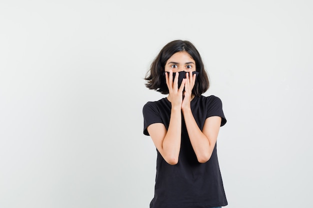 Foto gratuita bambina che tiene le mani sul viso in maglietta nera, maschera e guardando stupito, vista frontale.