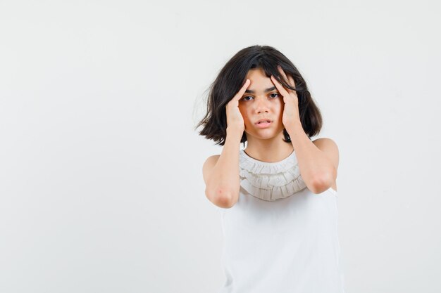 Little girl holding hands to head in white blouse and looking distressed , front view.