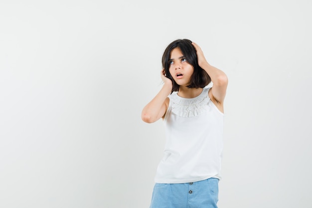 Little girl holding hands on head in white blouse, shorts and looking pensive , front view.