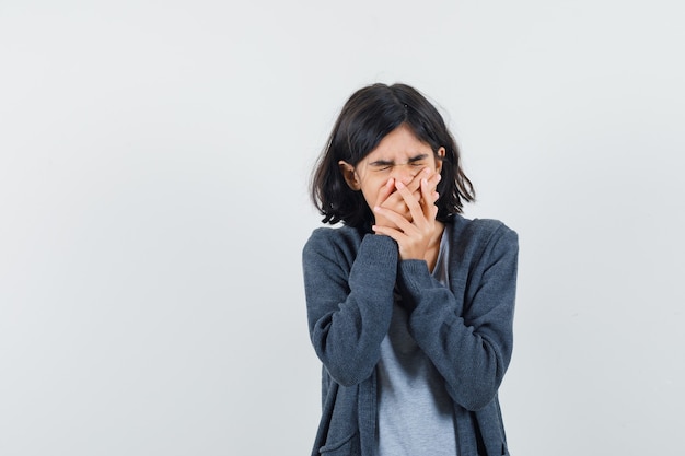 Little girl holding hands on mouth in t-shirt, jacket and looking mournful , front view.