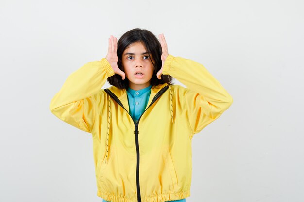 Little girl holding hands near sides of face in shirt, jacket and looking puzzled. front view.