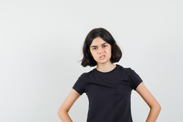 Little girl holding hands on waist in black t-shirt and looking angry. front view.