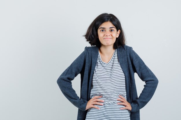 Little girl holding hands on waist in t-shirt, jacket and looking merry ,