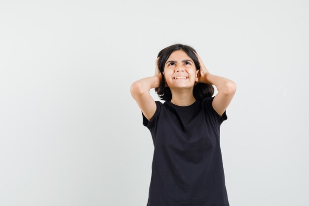 Маленькая девочка держит голову руками в черной футболке и смотрит с тревогой, вид спереди.