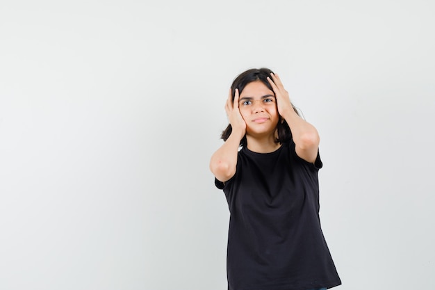 Little girl holding head with hands in black t-shirt and looking confused. front view.