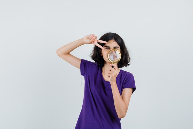 Little girl holding magnifying glass, showing v-sign in t-shirt ,