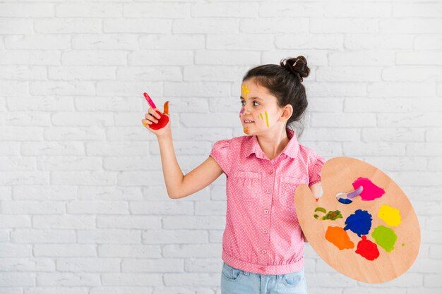 Little girl holding palette showing something with painted red finger